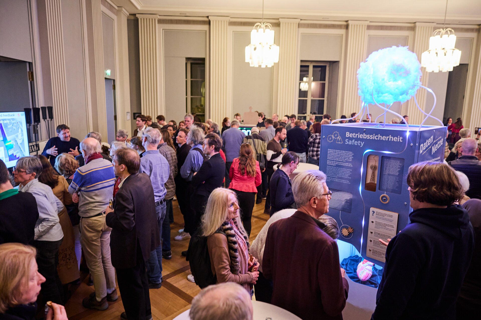 Crowd of people in a large room with chandeliers gathered around a blue display labeled 'Betriebssicherheit Safety'