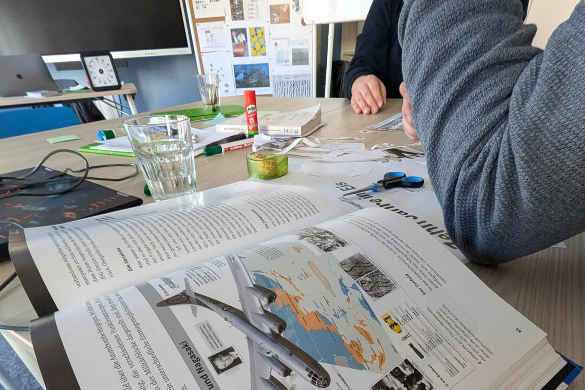 Two people working at a table with an open book on visualization. Visible items include a world map, scissors, glue, a glass of water, and a computer in the background.