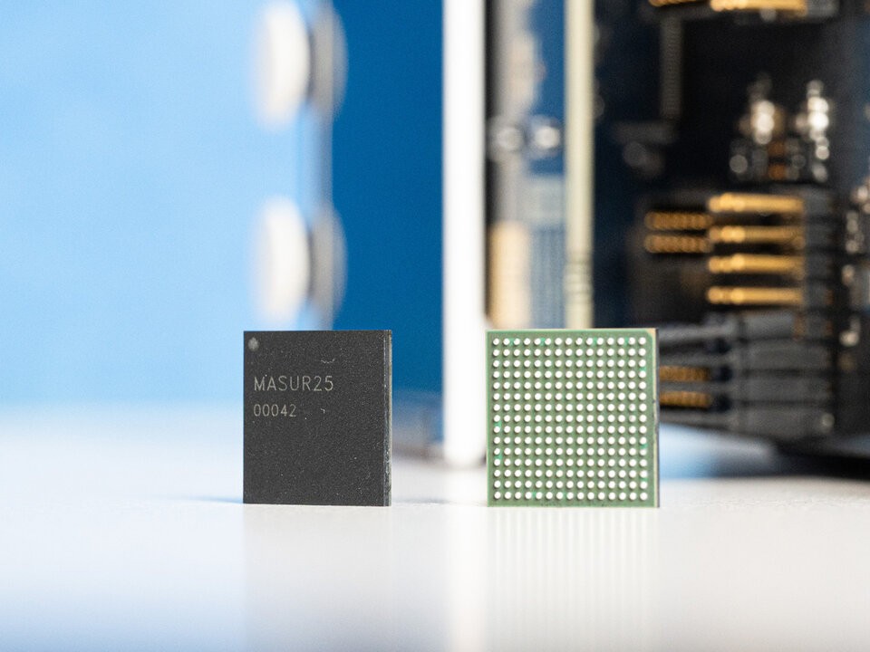 Zwei elektronische Bauteile stehen nebeneinander auf einer hellen Oberfläche. Links befindet sich ein quadratisches Chip Case mit matter, dunkler Oberfläche und der gut lesbaren Aufschrift 'MASUR25 00042'. Rechts daneben steht ein weiterer quadratischer Chip, dessen Oberseite aus einem Raster feiner, heller Kontaktpunkte besteht. Im Hintergrund sind unscharf Teile einer elektronischen Platine mit goldfarbenen Kontakten und blauer Fläche zu erkennen.