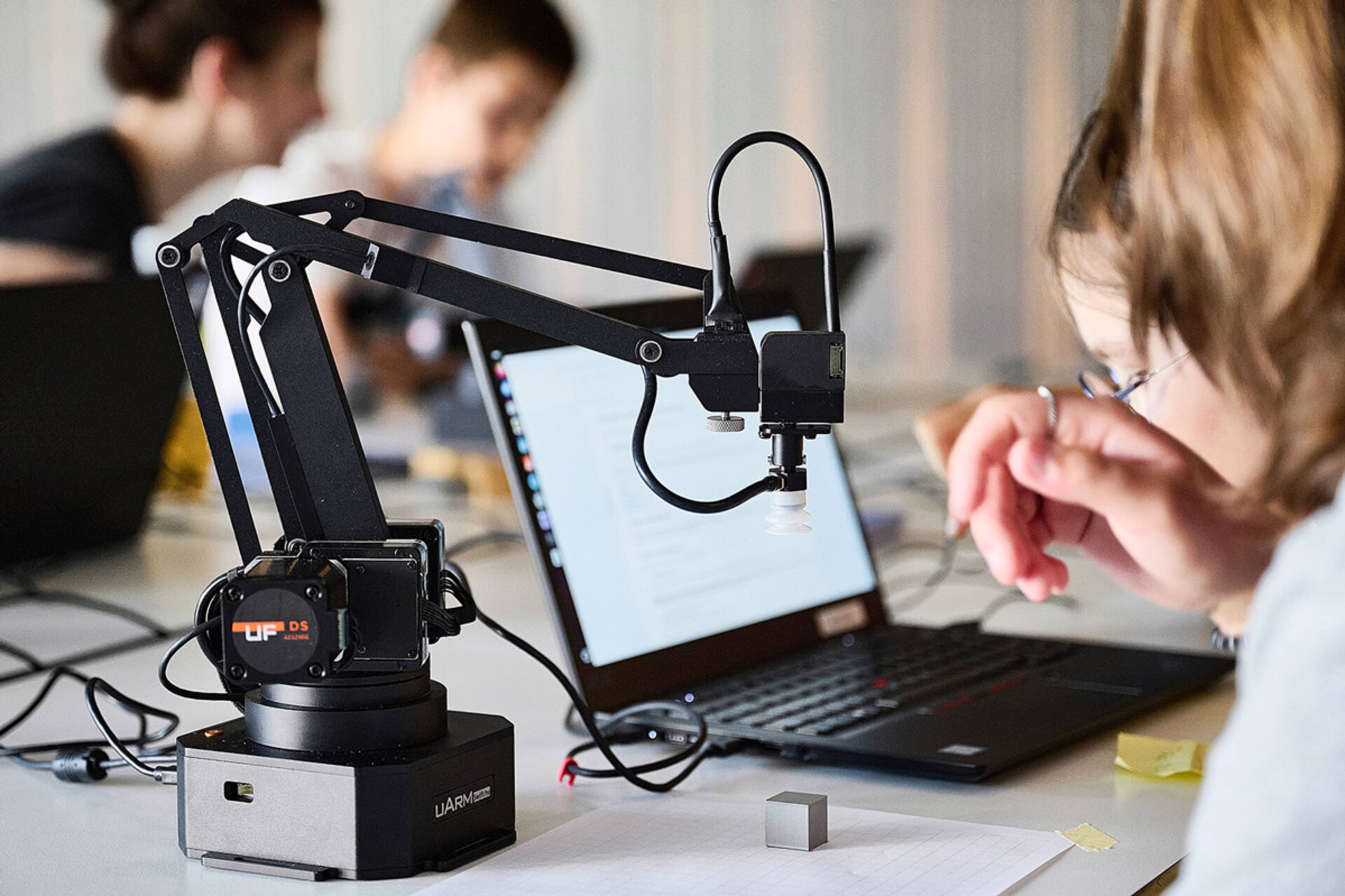 Roboterarm mit Kamera vor Laptop, im Hintergrund unscharfe Personen