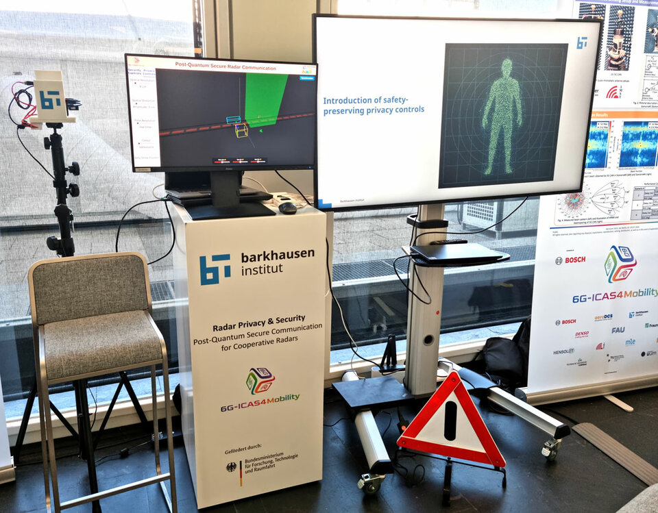 Exhibition booth with two monitors showing radar security and privacy controls, a tall chair, and a warning triangle sign on the floor.