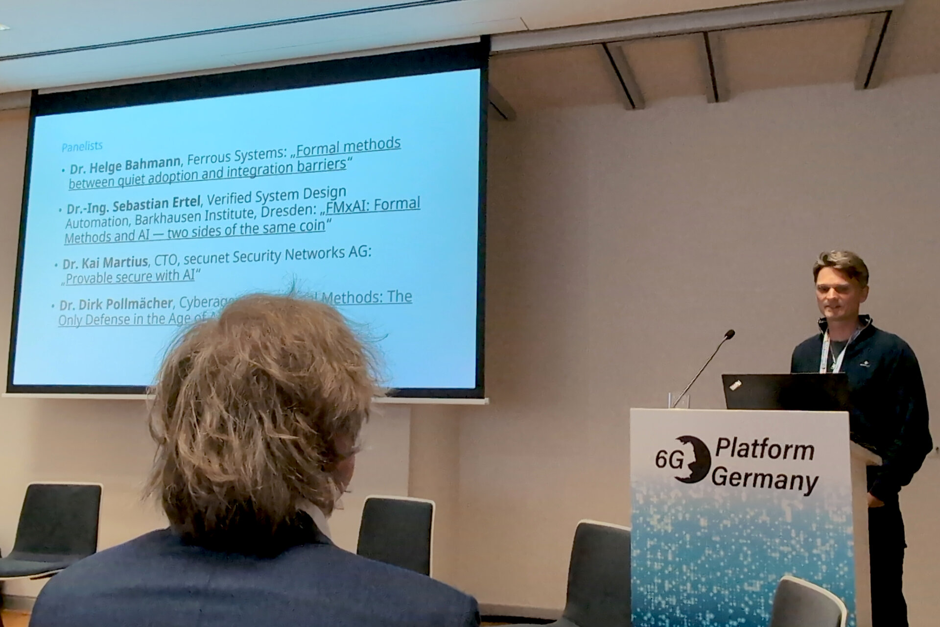 Man with short hair standing at a podium labeled '6G Platform Germany' giving a presentation to an audience member with gray hair in a conference room