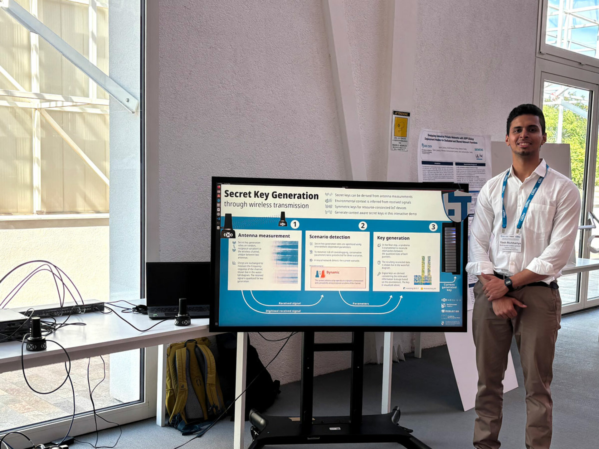 Ein Mann in einem weißen Hemd, mit einem Lanyard um den Hals und einem Namensschild mit der Aufschrift 'Yash Richhariya', steht neben einem großen Monitor, auf dem eine Präsentation mit dem Titel 'Secret Key Generation through wireless transmission' mit Diagrammen und Text angezeigt wird.