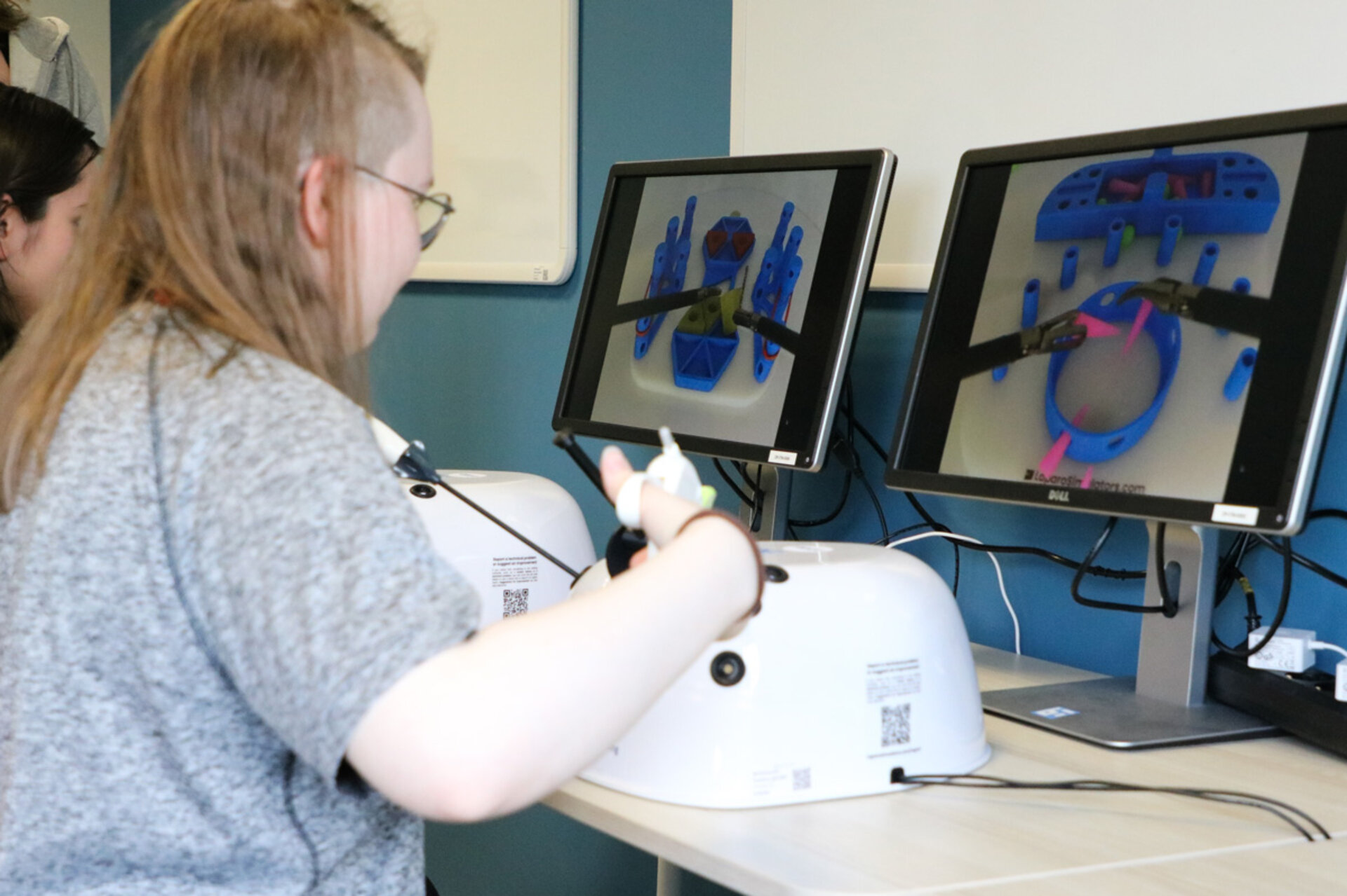 A person with glasses practices on a surgical simulator, using controls on a desk. Two monitors display colorful virtual surgical tools.