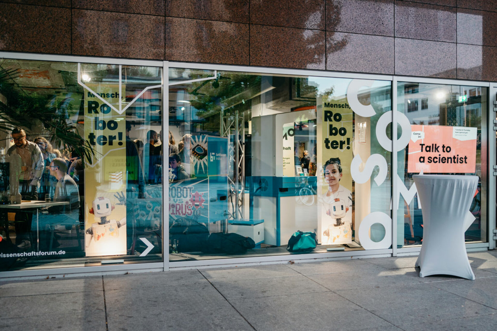 Glass facade with posters for the exhibition 'Mensch, Roboter! – Robotik aus der Dresdner Wissenschaft' and a standing table with a sign 'Talk to a scientist'.