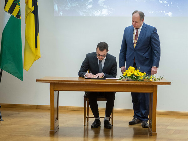 Andrey Ruzhanskiy sits at a table an signs a certificate. Mayor Hilbert stands beside him.