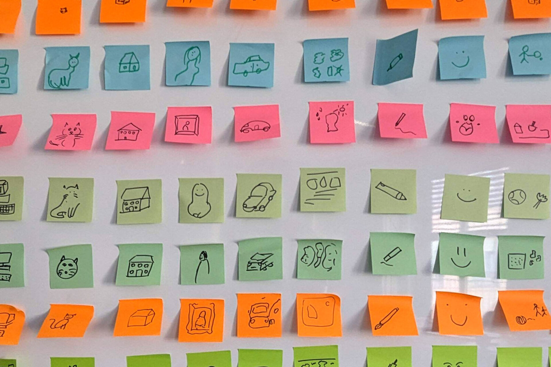 A wall covered with colorful sticky notes in green, orange, and blue, featuring simple drawings of animals, houses, vehicles, and other objects.