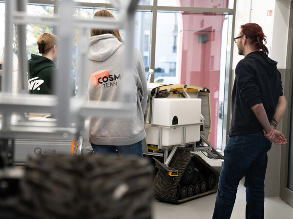 The picture shows three people looking at a small, chain-driven vehicle equipped with a white container. Two young women are standing facing the vehicle, one is wearing a gray sweatshirt with “COSMO TEAM” printed on it. A man with red hair stands with his back to the vehicle and looks at it from the side. The background consists of a bright, modern room with large windows. The focus is on the vehicle and the people, the room is blurred.