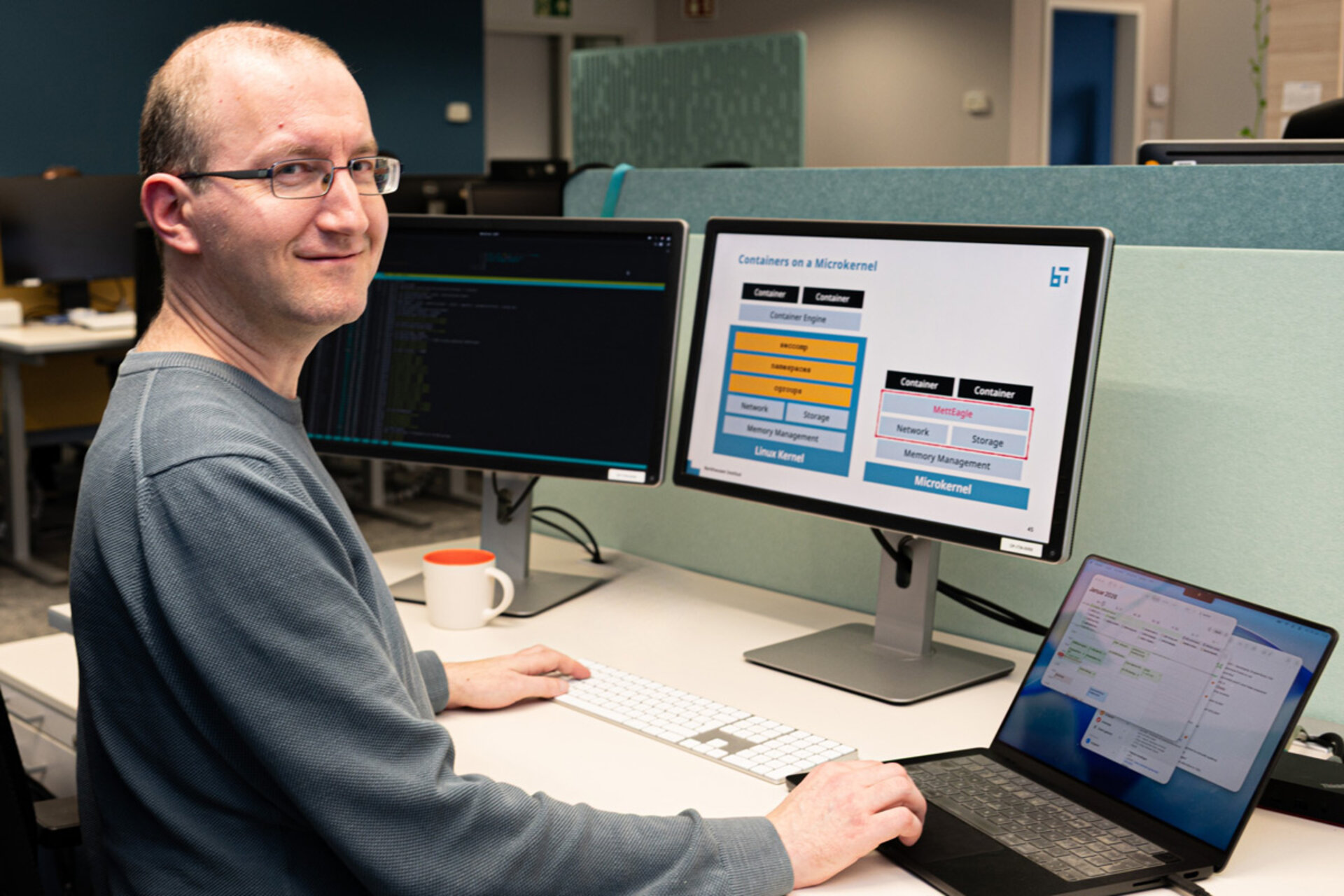 Person sitzt an einem Schreibtisch vor zwei Monitoren und einem Laptop, auf einem Monitor ist eine Präsentation zu Containern und Microkernel zu sehen
