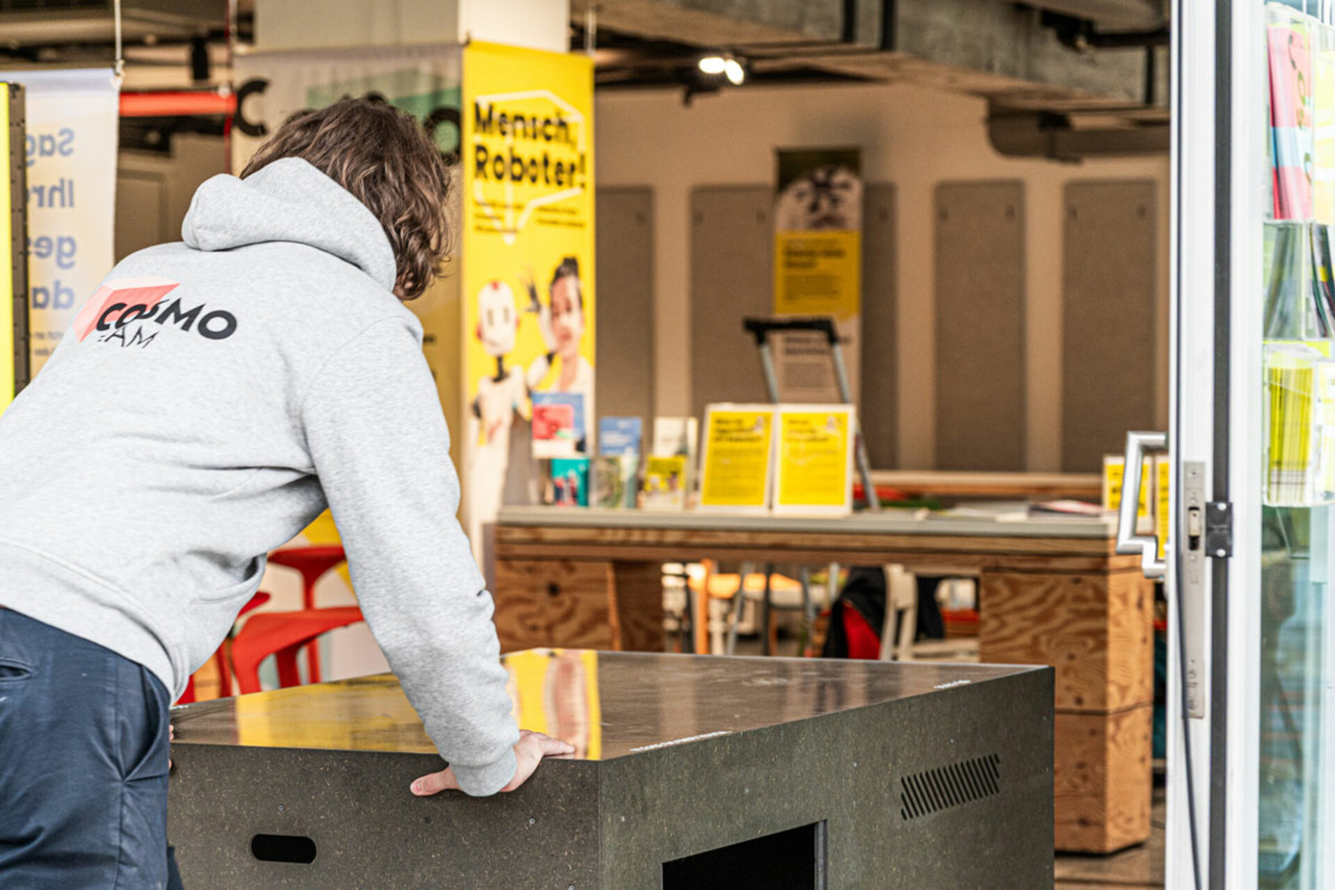 Person mit grauem COSMO-Hoodie schiebt eine schwarze Kiste in den Ausstellungsraum der Ausstellung 'Mensch, Roboter!' im COSMO Wissenschaftsforum.