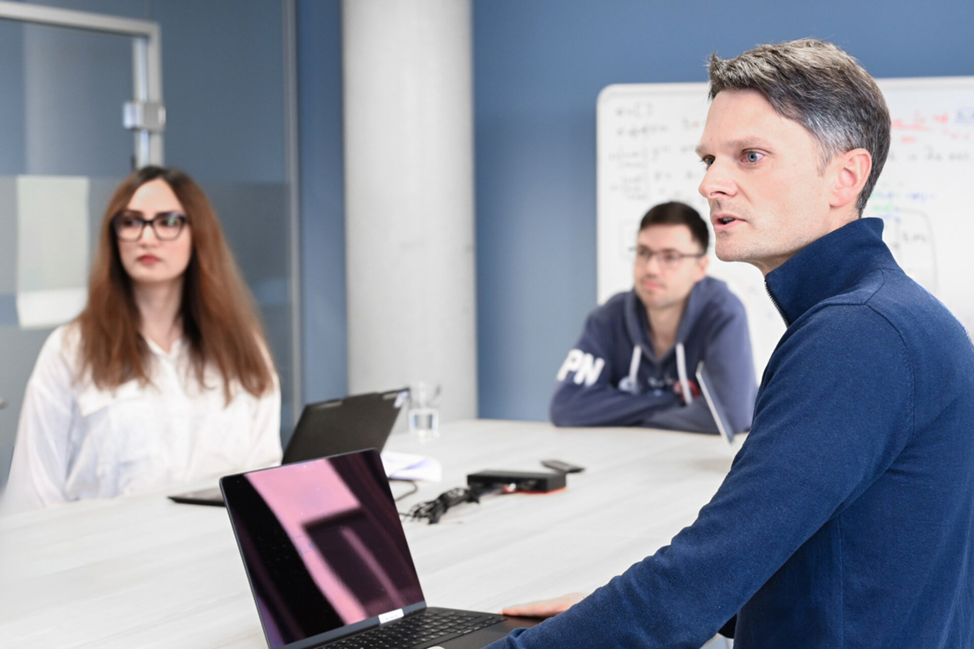Drei Personen sitzen an einem Tisch in einem modernen Besprechungsraum. Im Vordergrund ist eine Person mit kurzem, grauem Haar und dunkelblauem Pullover im Profil zu sehen, die an einem aufgeklappten Laptop arbeitet. Im Hintergrund sitzen zwei weitere Personen, eine mit langen, braunen Haaren und weißer Bluse, die andere mit dunklem Kapuzenpullover. Auf dem Tisch stehen Laptops, ein Glas Wasser und ein Smartphone. Im Hintergrund hängt ein Whiteboard an einer blauen Wand.