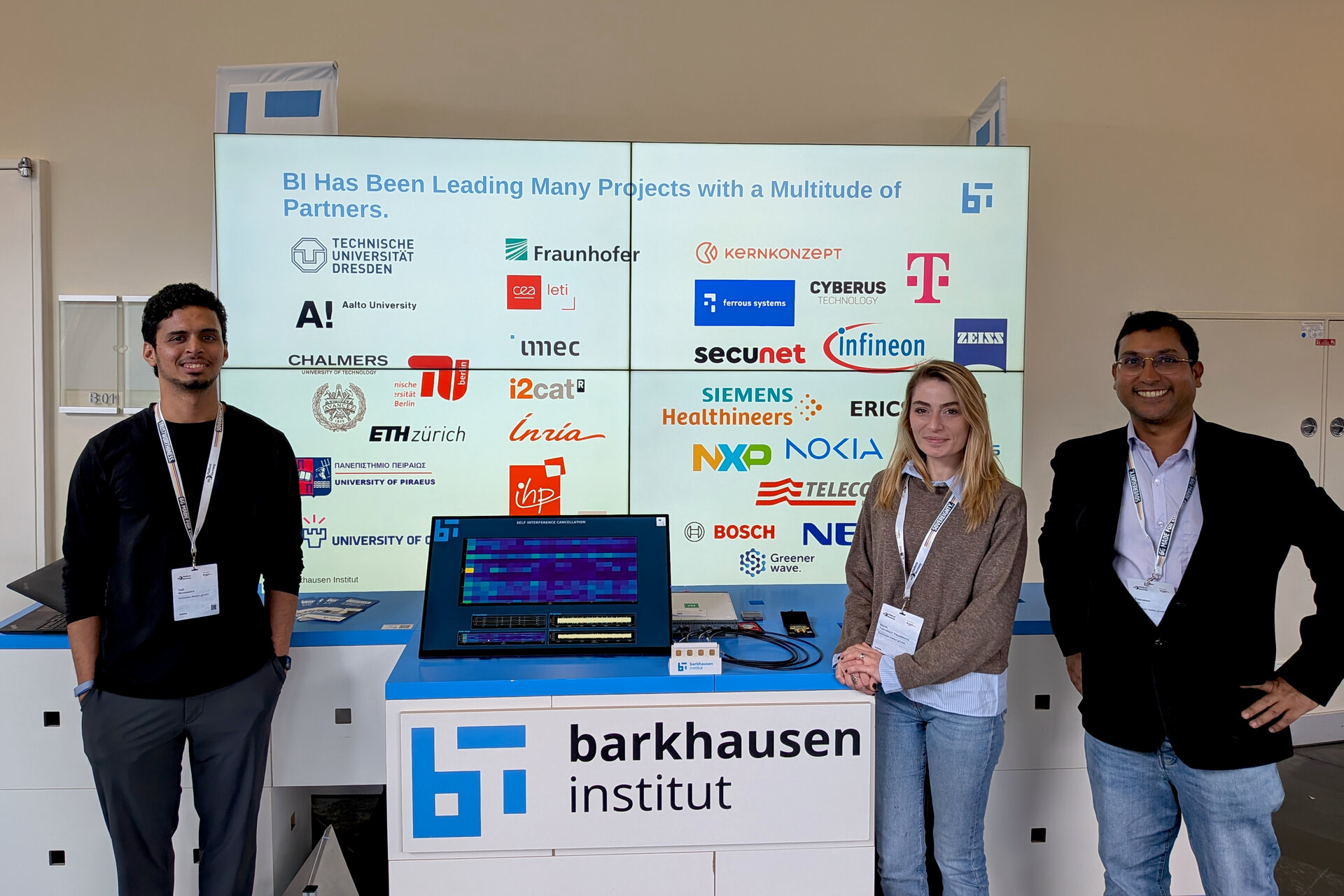 Three people standing behind a booth with a laptop and a sign reading 'barkhausen institut' and a screen behind showing logos of various partner organizations