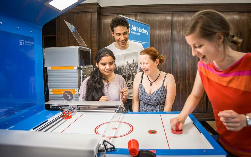 ein Mensch spielt am Airhockey Tisch. 3 weitere stehen daneben, lachen und haben Spaß.ckey Tisch, lachen und haben Spaß