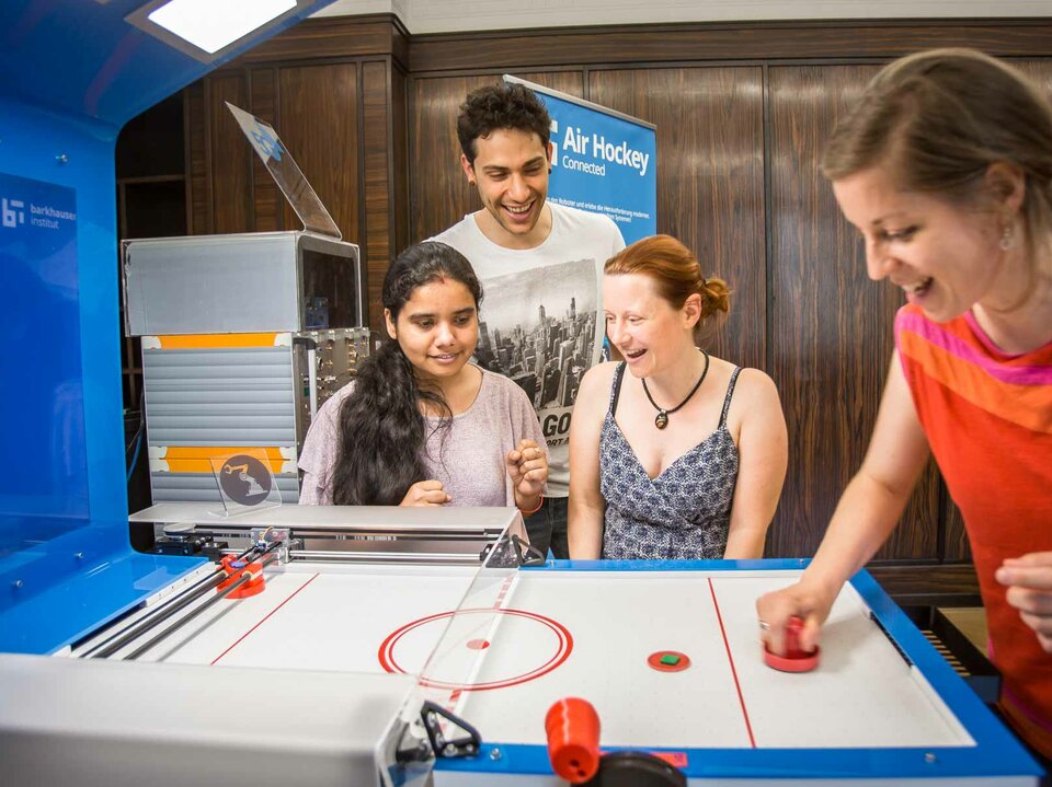 ein Mensch spielt am Airhockey Tisch. 3 weitere stehen daneben, lachen und haben Spaß.ckey Tisch, lachen und haben Spaß
