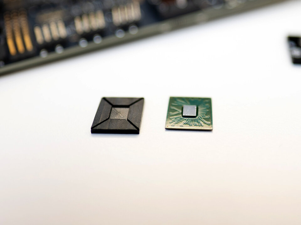 Zwei Halbleiterchips liegen nebeneinander auf einer hellen Oberfläche. Links befindet sich ein Chip in einem geschlossenen, dunklen Gehäuse mit abgeschrägten Kanten. Rechts liegt ein Chip mit sichtbaren Leiterbahnen und freiliegendem Siliziumkern. Im Hintergrund ist unscharf eine Leiterplatte zu erkennen. Das Bild illustriert den Verpackungsschritt, bei dem der Chip in ein schützendes Gehäuse eingebracht und die elektrischen Kontakte verbunden werden.