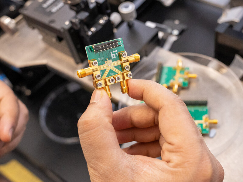 Hand holding a small green circuit board with gold connectors and screws in a laboratory setting