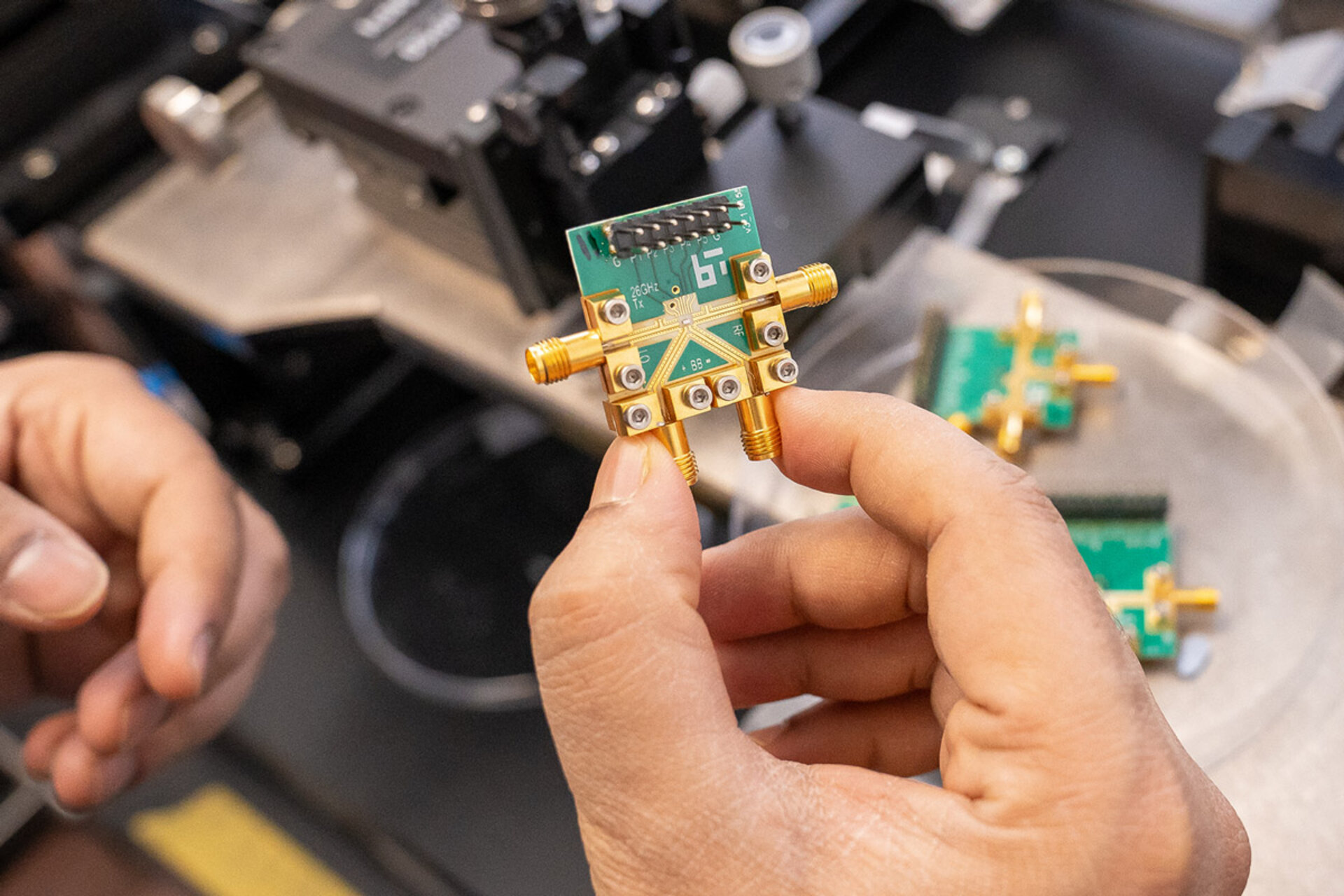 Hand holding a small green circuit board with gold connectors and screws in a laboratory setting