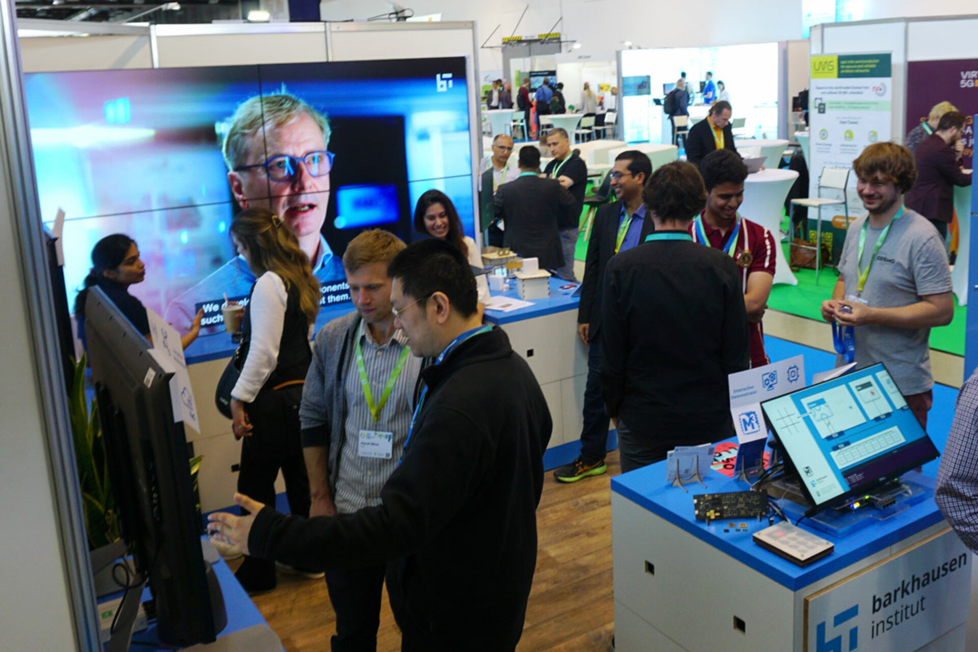 Menschen am Messestand des BI mit Bildschirmen und Informationsmaterial auf dem 6G Summit in der Messehalle Dresden