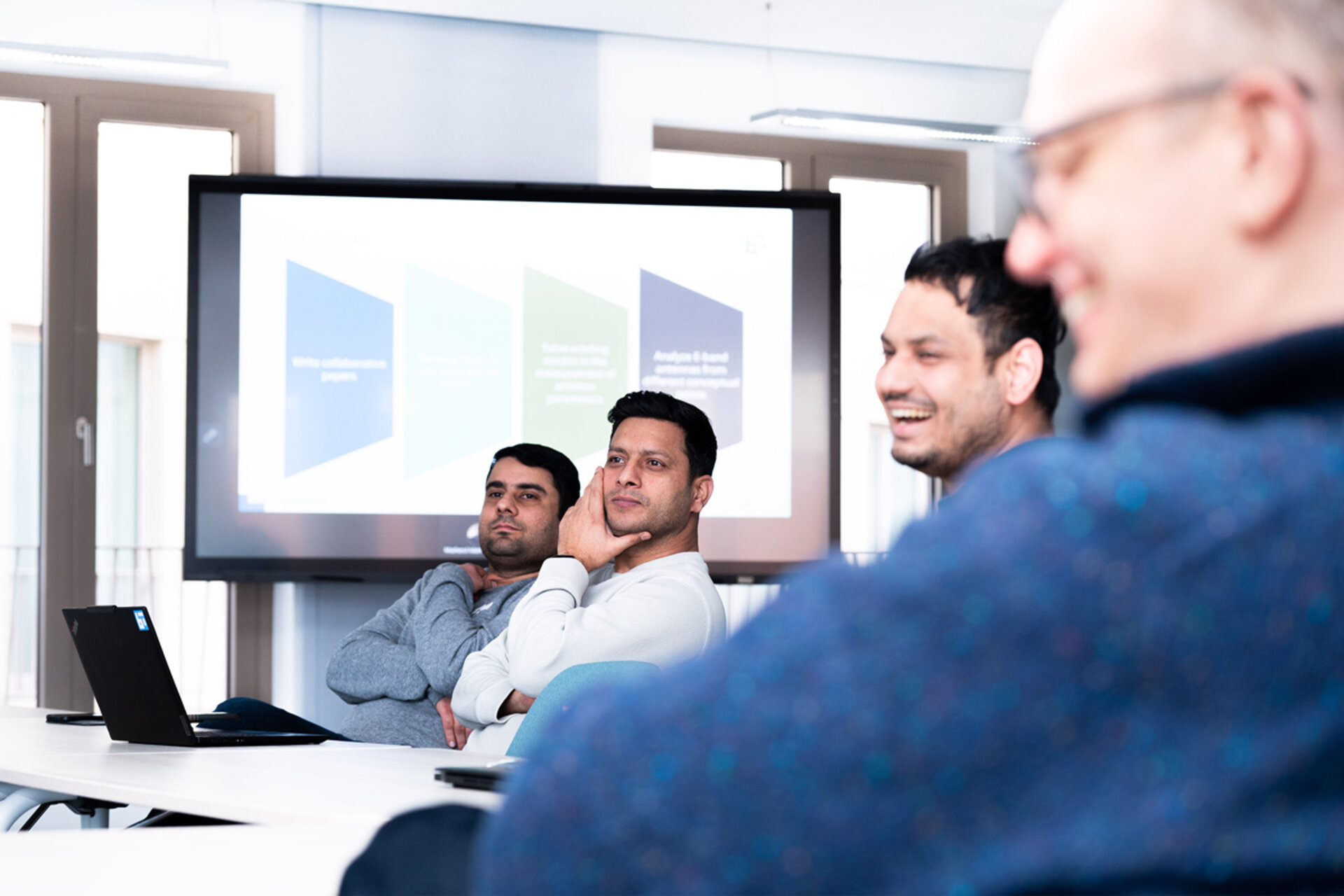 Three people sitting at a table in a meeting room with a presentation screen in the background