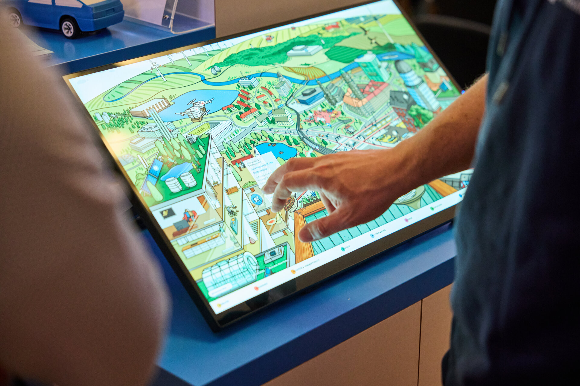 Person pointing at a colorful illustrated map on a horizontal touchscreen display with another person nearby