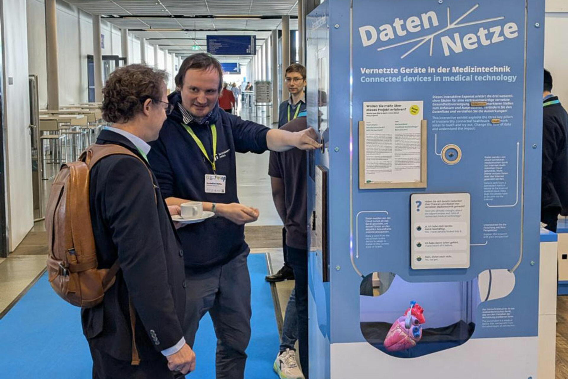 Zwei Männer im Gespräch vor einem blauen Informationsstand mit dem Titel 'Daten Netze' über vernetzte Geräte in der Medizintechnik, darunter ein Modell eines Herzens.