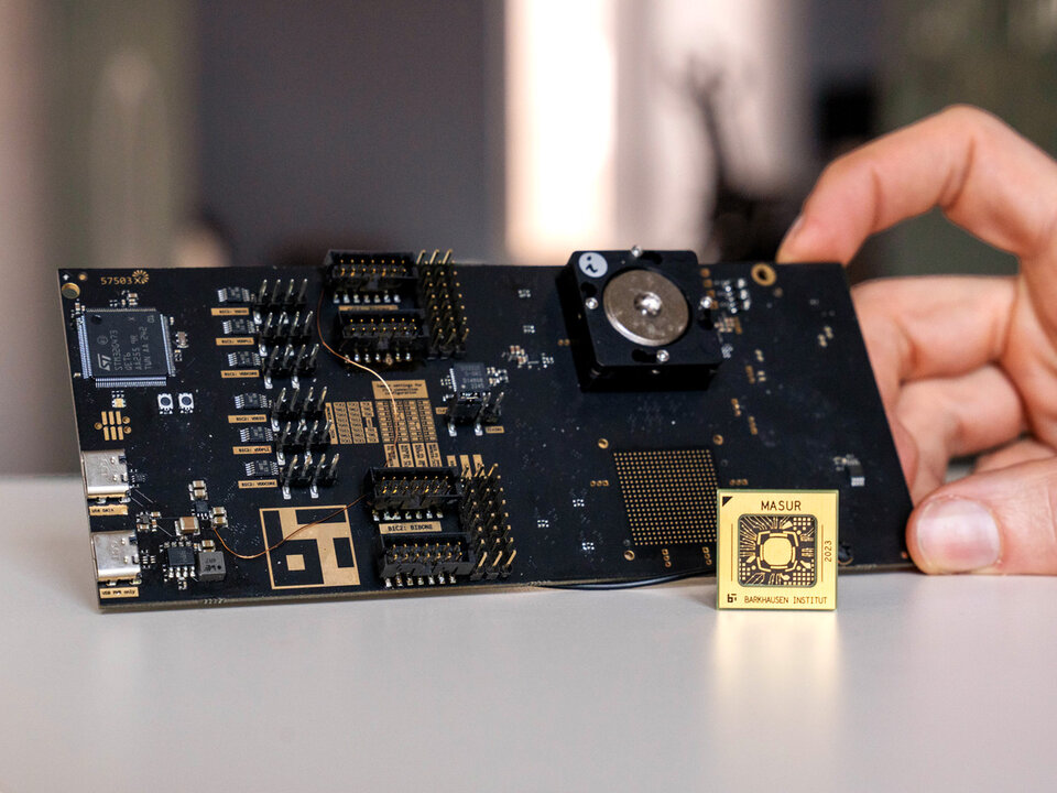 Eine Hand hält eine schwarze Leiterplatte mit mehreren Anschlüssen und Bauteilen, daneben ein kleiner goldfarbener Plattformchip mit der Aufschrift MASUR 2023" vom Barkhausen Institut