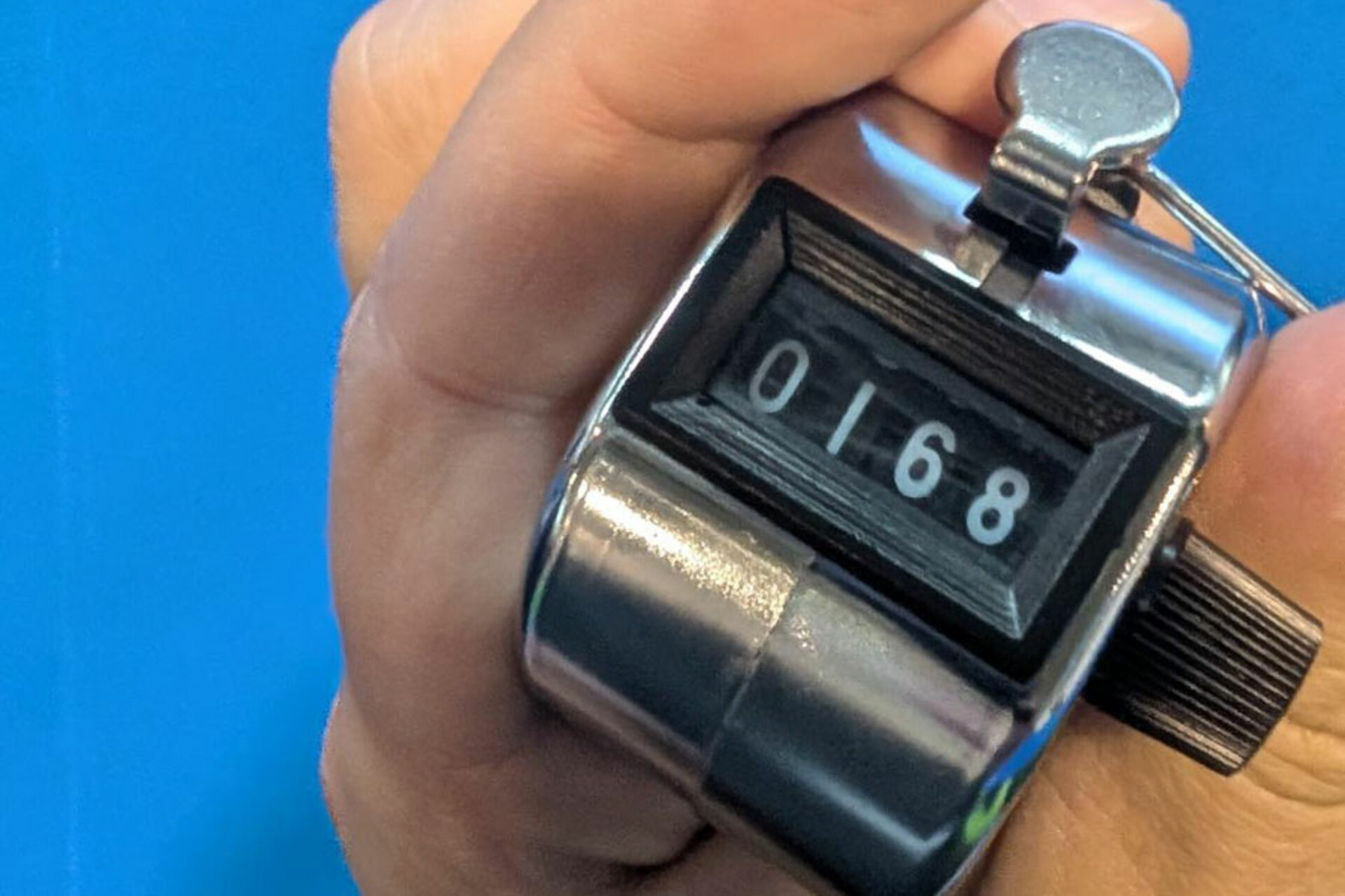 Hand hält einen silbernen mechanischen Zähler mit der Anzeige 0168 vor blauem Hintergrund.