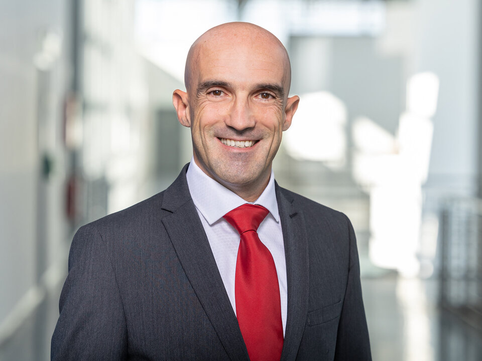 Mann in dunklem Anzug mit weißem Hemd und roter Krawatte steht in einem hellen Flur mit Glaswänden.