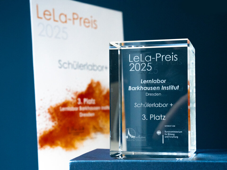 Auf dem Bild ist ein kleiner Glaskubus zu sehen. Darauf steht "LeLa-Preis 2025. Lernlabor Barkhausen Institut Dresden. Schülerlabor+. 3. Platz. Im Hintergrund ist eine Urkunde in weiß-orange mit dem gleichen Text leicht verschwommen zu erkennen.