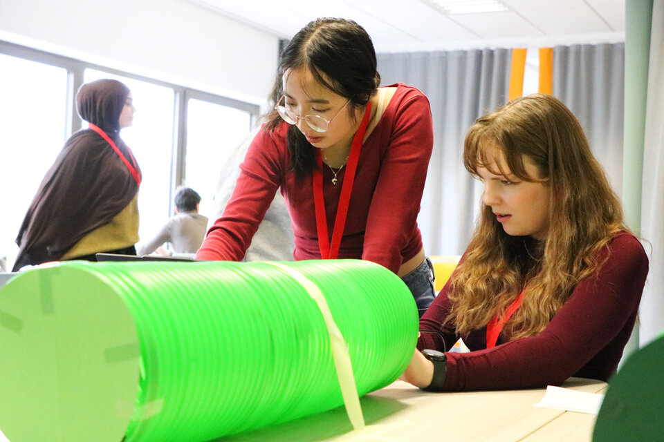Two women focus on assembling a project with a large green tube at a table. They wear red lanyards. Others interact in the background, creating a collaborative atmosphere.