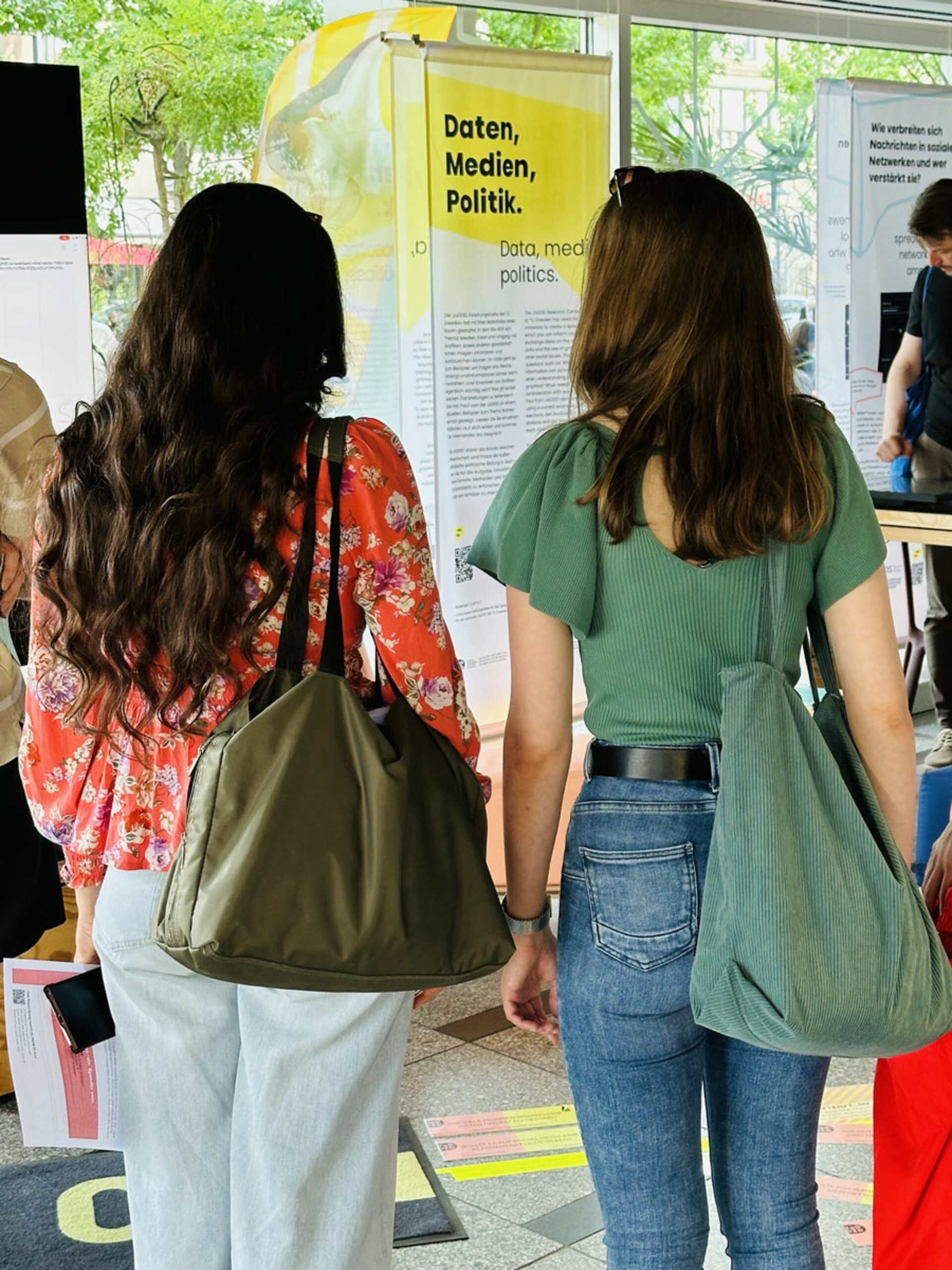 Zwei Frauen mit Taschen stehen vor einer Ausstellung mit dem Titel „Daten, Medien, Politik“. Die Szenerie erinnert an eine moderne Ausstellung oder öffentliche Veranstaltung.