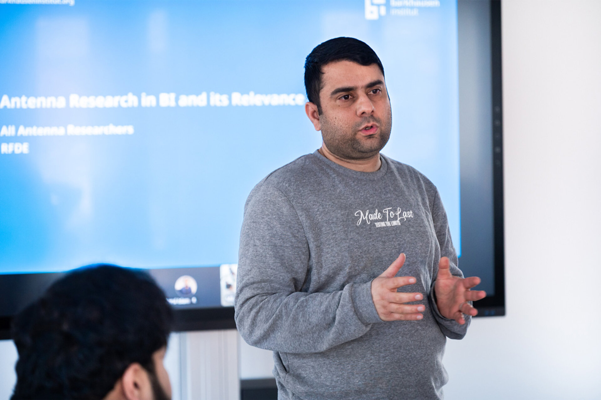 Man in gray sweatshirt giving a presentation in front of a screen with text