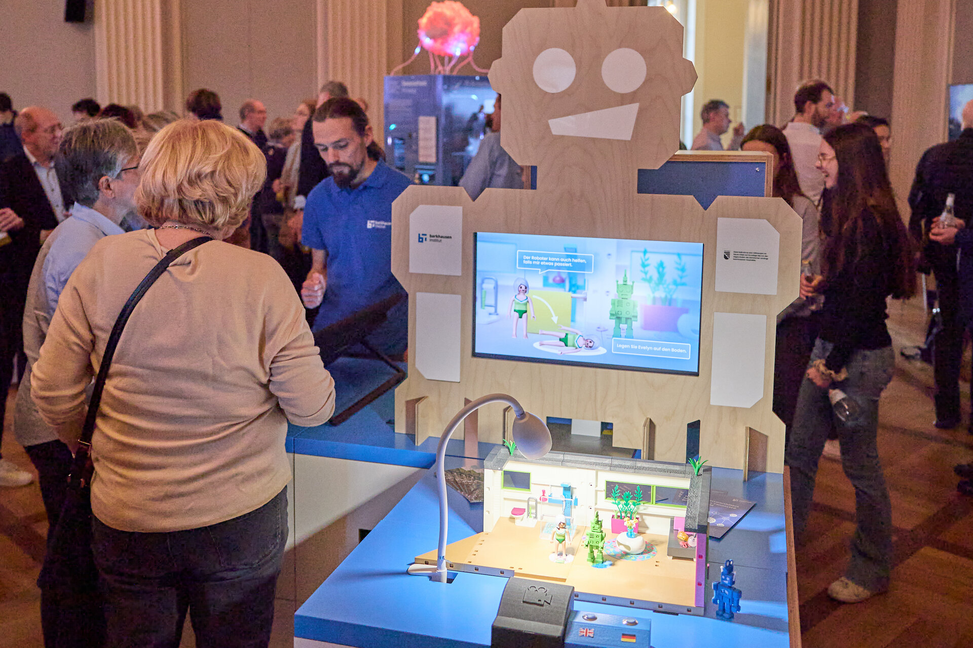Exhibition room with people around a robot-shaped display featuring a screen and a miniature model on a blue table