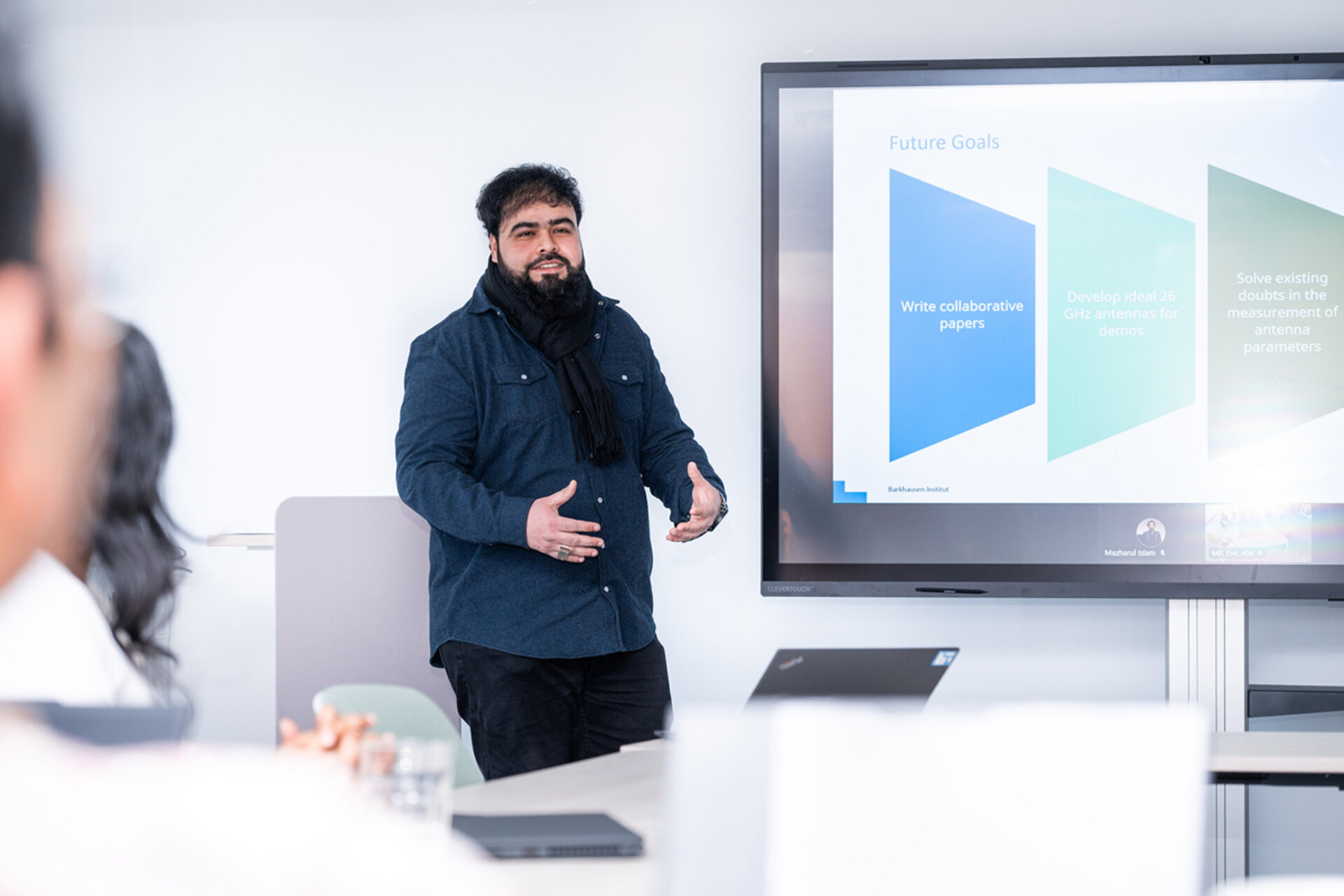 Man standing and gesturing in front of a screen showing a presentation slide titled 'Future Goals' in a meeting room