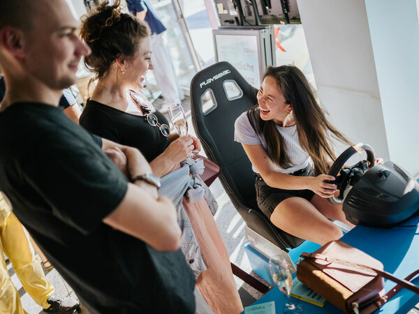 A woman sits at the steering wheel of a driving simulator, another woman watches, both laugh.