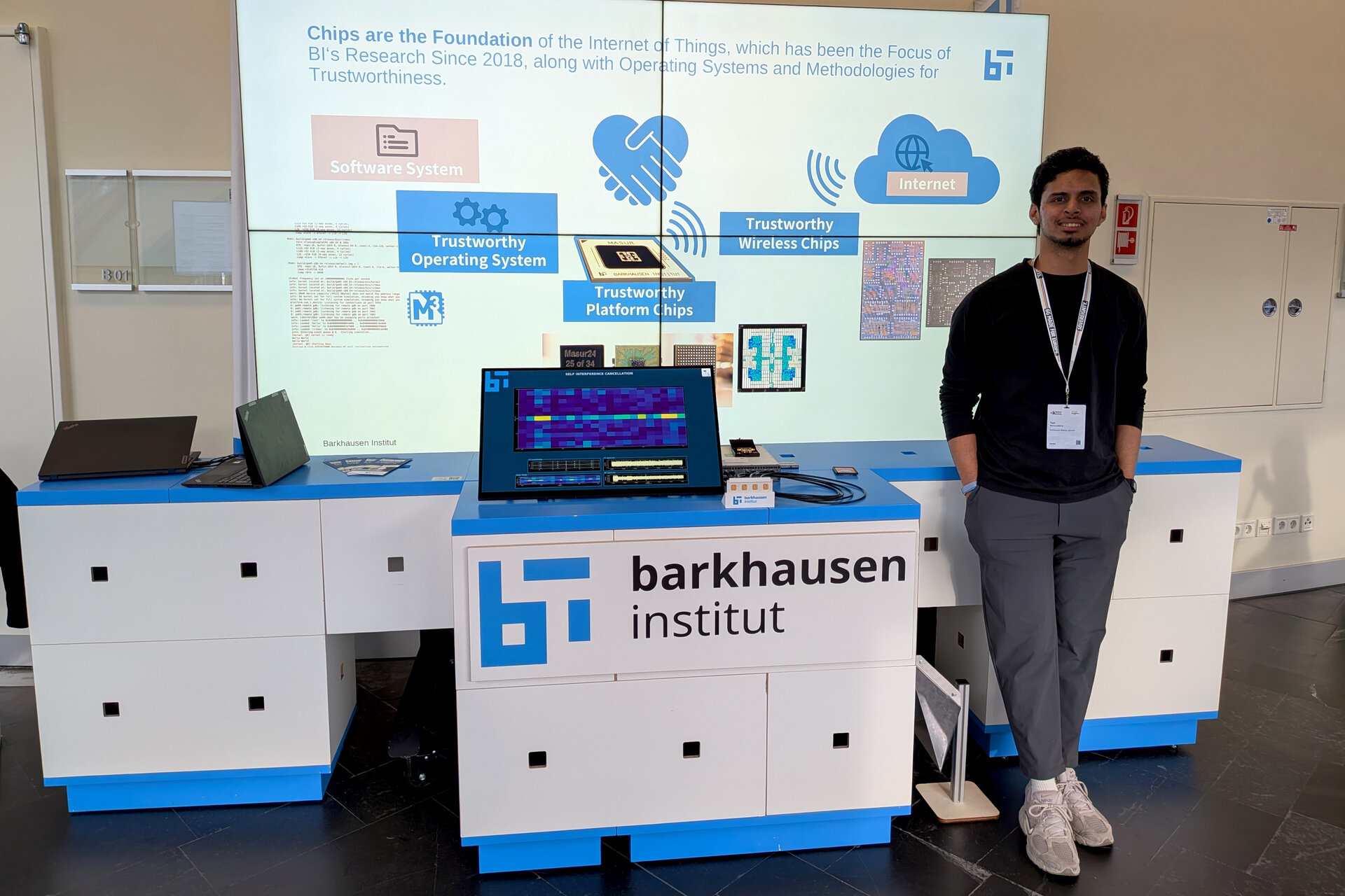 Man standing next to a booth with a laptop and a monitor displaying a colorful chart, booth labeled 'barkhausen institut' with a white and blue design and a backdrop showing a diagram about trustworthy chips and operating Systems