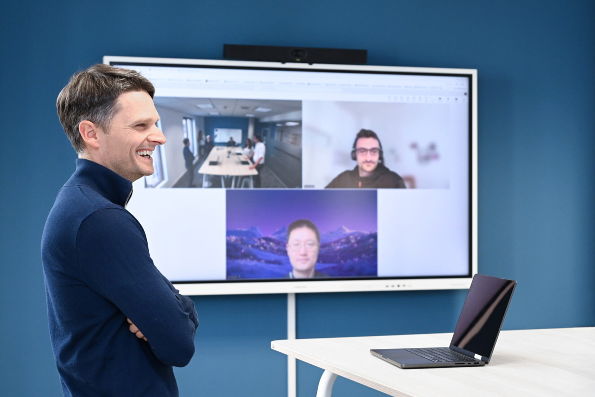 Person mit verschränkten Armen steht vor einem großen Bildschirm, auf dem eine Videokonferenz mit mehreren Teilnehmern zu sehen ist, daneben steht ein Laptop auf einem Tisch