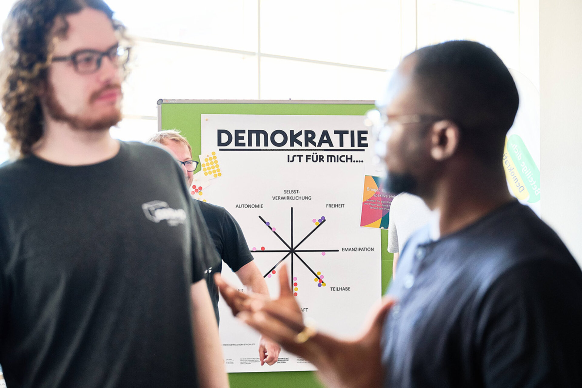 Zwei Männer im Gespräch vor einem Plakat mit dem Titel 'DEMOKRATIE IST FÜR MICH....' und einem Diagramm mit Begriffen wie 'AUTONOMIE', 'FREIHEIT' und 'TEILHABE'.