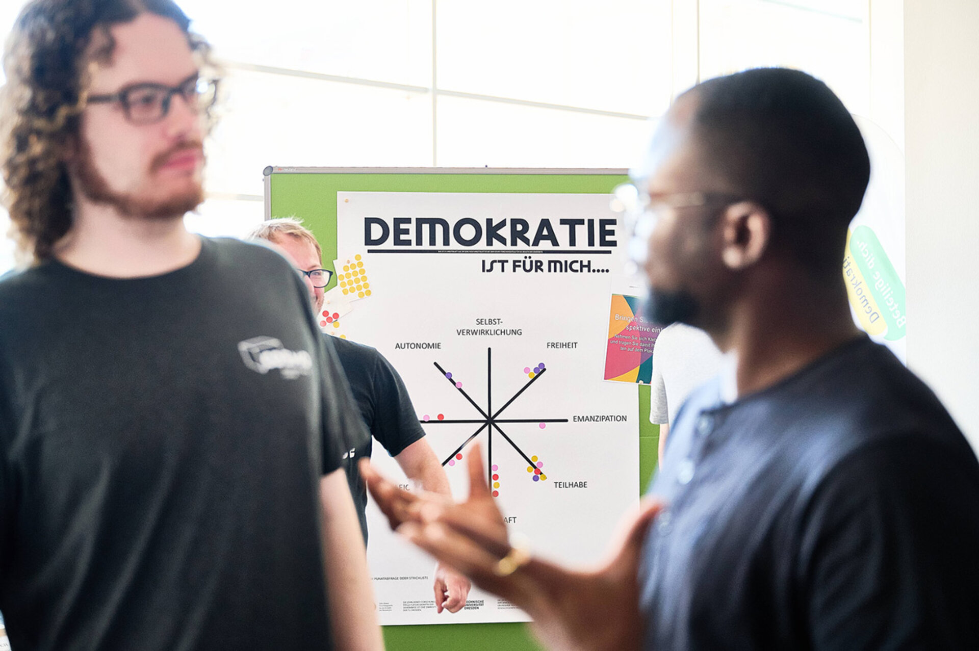 Zwei Männer im Gespräch vor einem Plakat mit dem Titel 'DEMOKRATIE IST FÜR MICH....' und einem Diagramm mit Begriffen wie 'AUTONOMIE', 'FREIHEIT' und 'TEILHABE'.