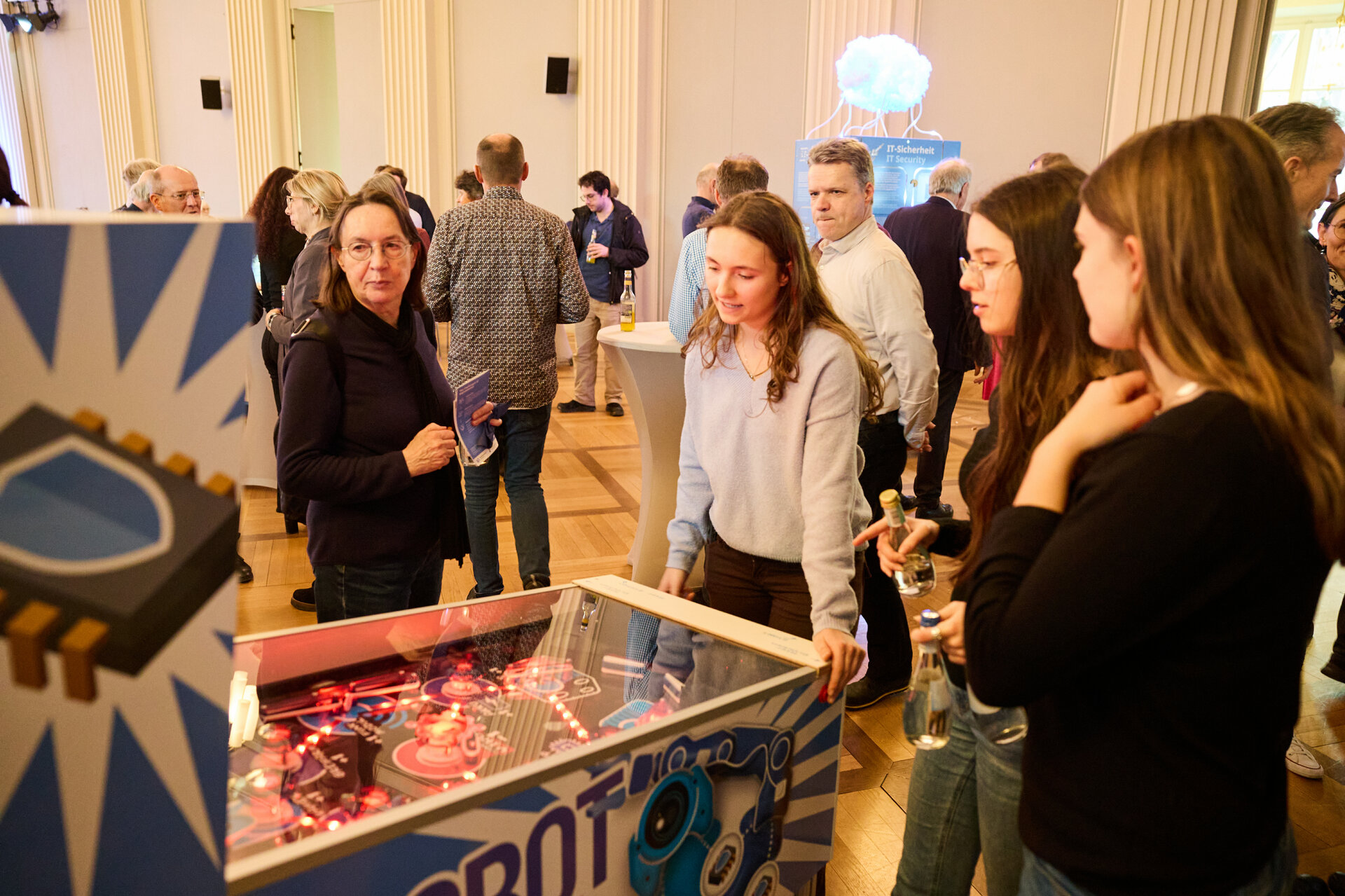 Group of people gathered around a pinball machine in a large room with columns