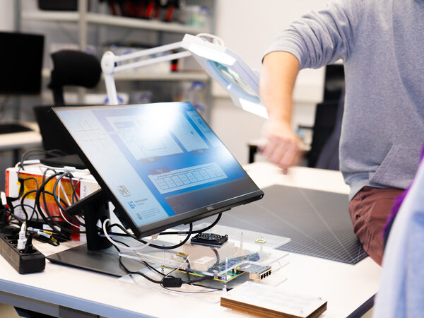The picture shows a workstation in a laboratory or workshop at the Barkhausen Institute. In the foreground is a computer monitor on which a program with graphical representations can be seen. Surrounding the monitor are various cables and electronic components, which are mounted on a table with a self-made, transparent holder. Part of a person can be seen in the background, apparently working on setting up or operating the equipment. The atmosphere is technical and concentrated.