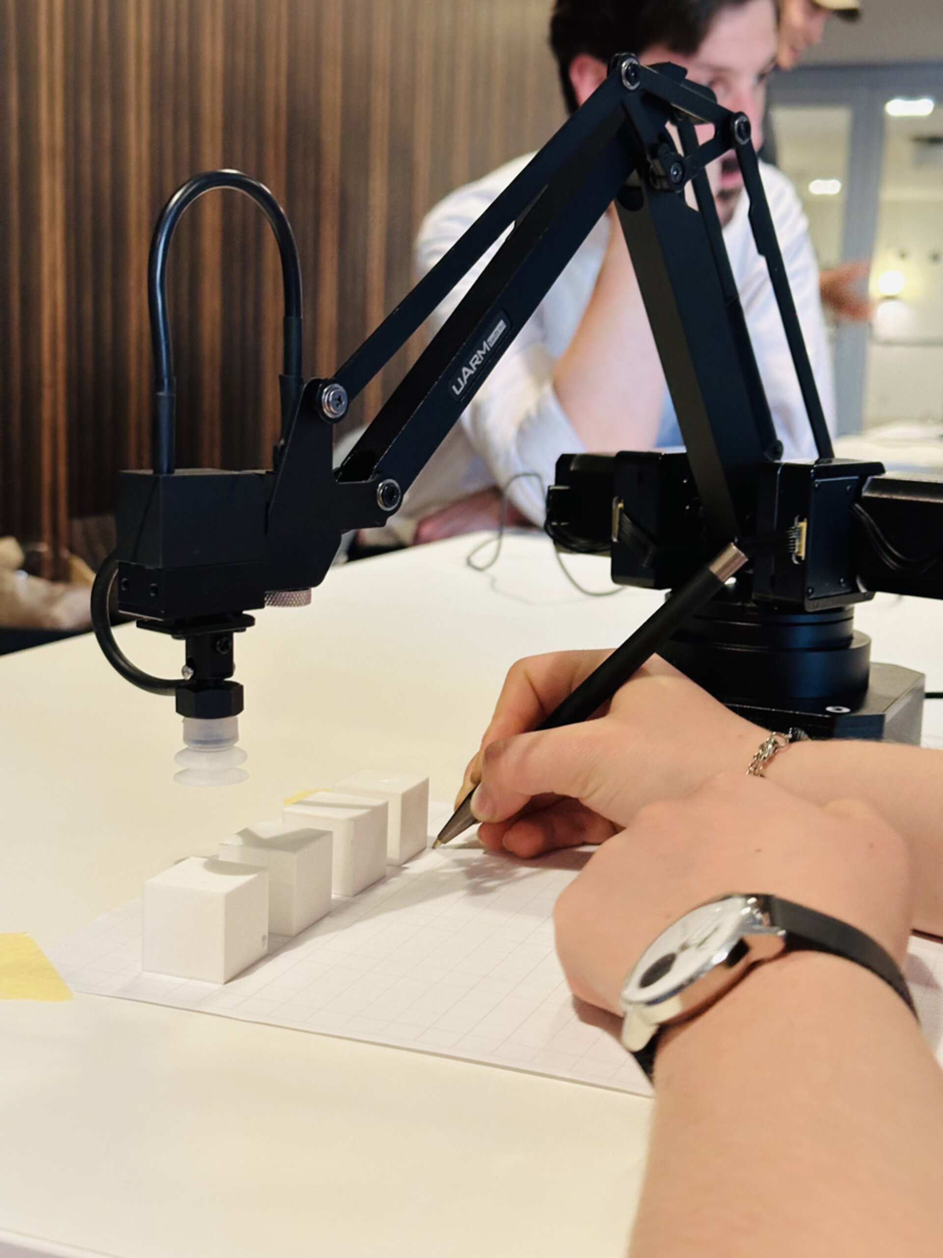 Ein Roboterarm und eine Person an einem Tisch interagieren mit kleinen weißen Blöcken. Die Umgebung wirkt konzentriert und engagiert.