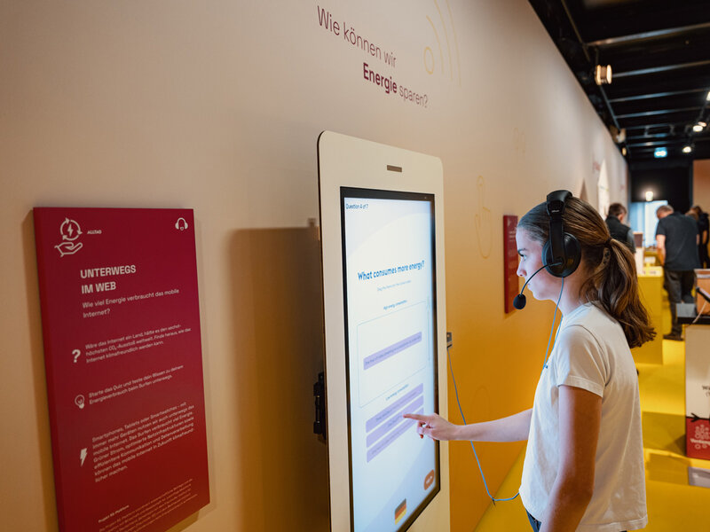Person mit Kopfhörern bedient interaktiven Touchscreen zum Thema Energieverbrauch im Web bei Ausstellung auf der MS Wissenschaft.