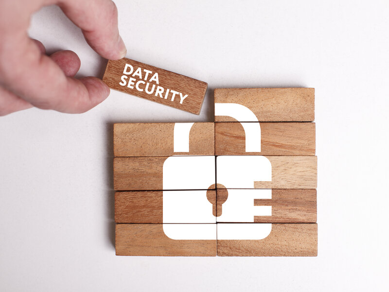 There are wooden blocks assembled into a rectangle. Folded together shows a white printed image of a lock with a keyhole. The upper left corner of the rectangle is just put down. The fingers of the hand doing this can be seen. On the cornerstone is written in white letters "Data Security".