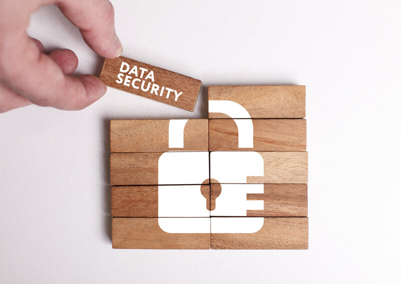 There are wooden blocks assembled into a rectangle. Folded together shows a white printed image of a lock with a keyhole. The upper left corner of the rectangle is just put down. The fingers of the hand doing this can be seen. On the cornerstone is written in white letters "Data Security".