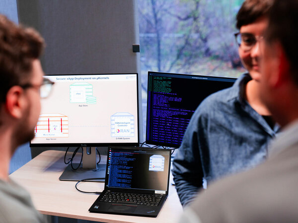 Eine Gruppe von Personen diskutiert vor zwei Monitoren. Einer zeigt eine Grafik zur sicheren Bereitstellung von xApps, während der andere Programmiercode anzeigt. Auf dem Tisch steht ein Laptop, der mit den Monitoren verbunden ist. Im Hintergrund sieht man ein Fenster mit Blick ins Freie.