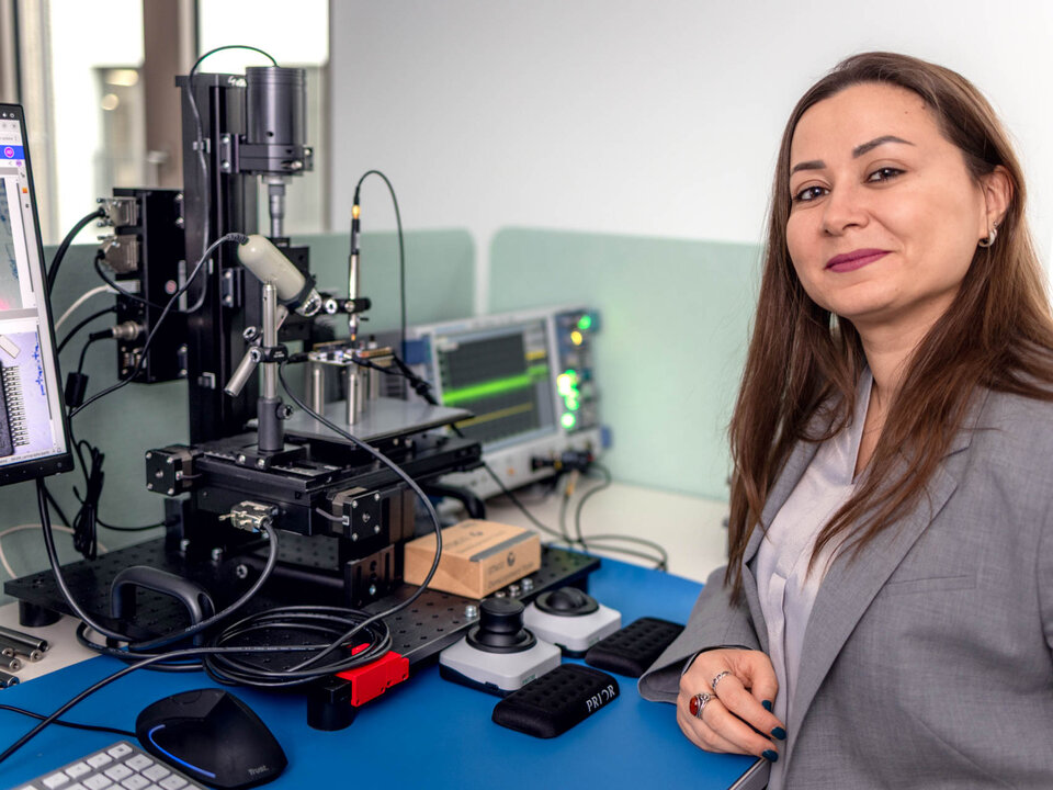 Prof. Elif Bilge Kavun trägt einen hellgrauen Blazer und steht an einem blauen Labortisch. Auf dem Tisch befinden sich verschiedene Laborgeräte, darunter ein Mikroskop mit mehreren Kabeln und Sensoren, ein Monitor mit Messkurven, eine Computermaus, ein Tastaturfeld und mehrere elektronische Komponenten. Die Umgebung wirkt wie ein moderner Laborarbeitsplatz.
