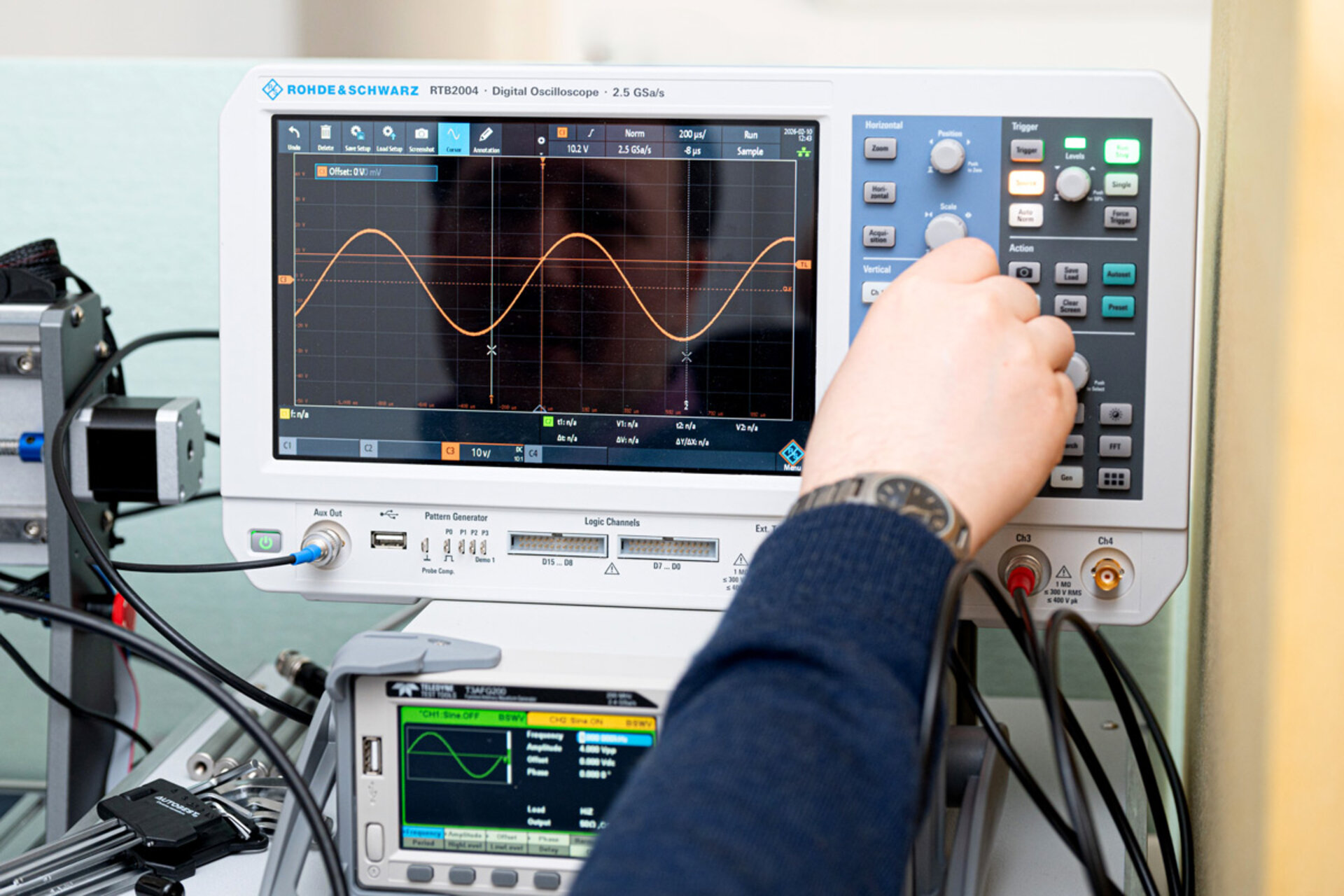 Hand adjusting controls on a Rohde & Schwarz digital oscilloscope displaying a sine wave