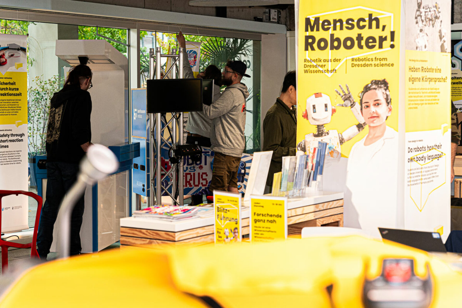 Gelber Ausstellungsbanner mit der Aufschrift 'Mensch, Roboter! Robotics from Dresden science' neben einem Tisch mit Flyern und Büchern, im Hintergrund Personen beim Aufbau der Ausstellung.