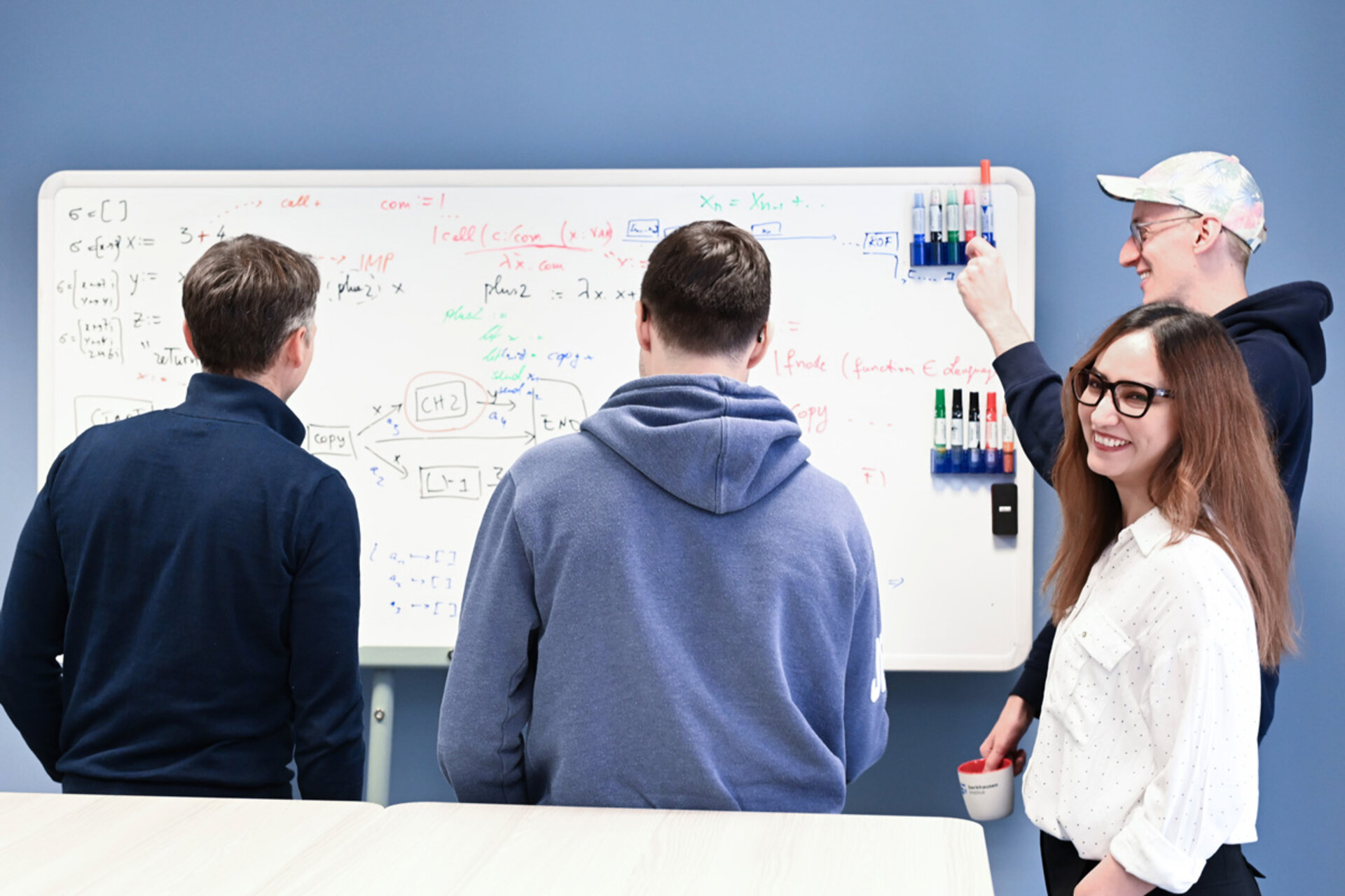 Vier Personen stehen vor einem Whiteboard mit mathematischen Formeln und Diagrammen, eine Person hält einen Marker in der Hand